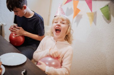 Kardeşi parti malzemeleri hazırlamaya odaklanırken genç bir kız elinde balonla gülerken neşeli bir kardeş anı başlıyor. Renkli dekorasyonlar şenlikli atmosfere ekleniyor.