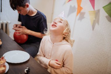 Genç bir kız parti şapkası takarken sevinçle gülerken, ağabeyi yakınlarda oturuyor ve kırmızı bir balon hazırlıyor. Renkli dekorasyonlar ve doğum günü hediyeleri masayı doldurur..