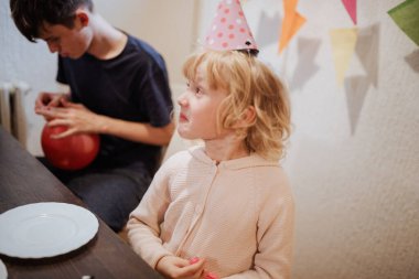 Parti şapkalı genç bir kız, kardeşinin kırmızı bir balon hazırlamasını heyecanla izliyor. Renkli dekorasyonlar odayı süslüyor, neşeli bir doğum günü havası yaratıyor..