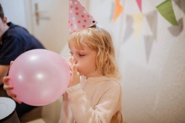Genç bir çocuk parti şapkası takıyor ve pembe bir balonu şenlikli renklerle dolu sıcak, dekore edilmiş bir yerde şişiriyor. Aile üyeleri kutlamanın tadını çıkarsın..