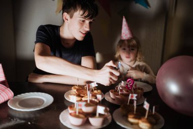 İki kardeş evde birlikte doğum günü kutlaması yapıyorlar. Büyük kardeş keklerin üzerine mum yakarken küçük kardeş hevesle etrafı şenlikli süslemeler ve balonlarla çevrili olarak izliyor..