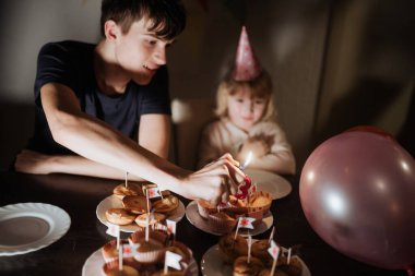 Genç bir çocuk kekin üzerine mum yakarken bir kız heyecanla izliyor. Parti şapkaları ve bayraklı kekler ev ortamında şenlikli bir ortam yaratıyor..