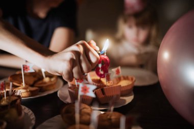Bir çocuk, büyük bir kardeşin doğum günü partisinde kekin üzerine mum yakmasını izliyor. Renkli dekorasyonlar ve onları çevreleyen neşeli bir atmosfer..