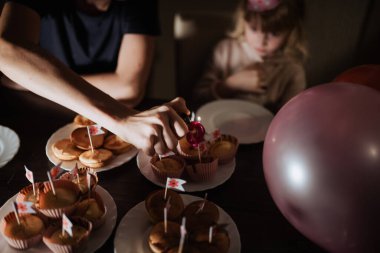Aile toplantısı sırasında, bir kardeş çörekleri mumlar ve bayraklarla süslerken, diğer kardeş de izliyor. Balonlar doğum günü kutlamasının neşeli atmosferine renk katar..
