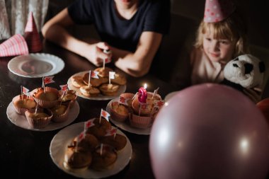 Küçük bayraklı kekler ve masada bir mum var. İki kardeş parti şapkası takıp, doğum günü partisinin rahat ortamının tadını çıkarırken kutlama yapıyorlar..