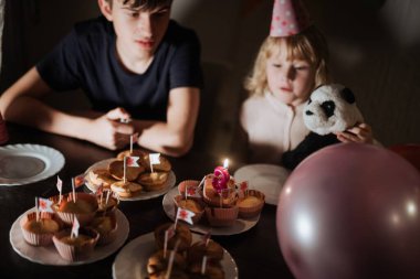 İki kardeş, evde bir masa dolusu süslü kek ve renkli balonlarla rahat bir doğum günü kutlamasının tadını çıkarıyorlar. Genç bir kız elinde bir panda oyuncağı tutuyor, anı güzelleştiriyor..