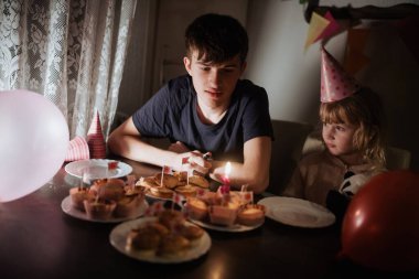 İki kardeş renkli kek ve balonlarla dolu bir doğum günü kutlamasının tadını çıkarıyorlar. Atmosfer şenlikli, parti şapkaları ve mum ışığı var..