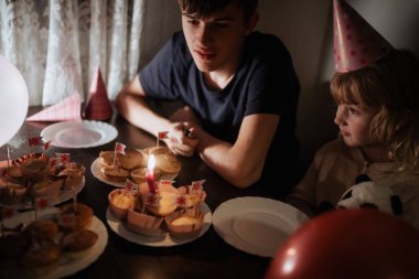 Kardeşler süslü çöreklerle dolu bir masada birlikte otururlar. Bir kardeş yanan bir mumu izlerken diğeri de kutlama sırasında sıcak ve şenlikli bir atmosfer yaratıyor..