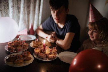 Genç bir erkek, bir keki mumla söndürmeye hazırlanan bir kızı gözlemler. Renkli süslemeler ve balonlar rahat evlerinde şenlikli bir atmosfer yaratıyor..