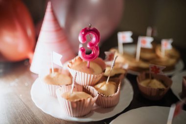 Masada üç numaralı pembe mumla, etrafı şenlikli süslemelerle çevrili lezzetli kekler sergileniyor. Balonlar ve parti şapkaları neşeli bir atmosfer yaratır.