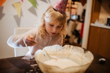 Parti şapkası takan bir çocuk hamuru bir kasede karıştırır, odaklanmış ve nişanlı. Renkli dekorasyonlar, aile üyelerinin kutlamaya hazırlandığı şenlikli bir ortam yaratıyor..