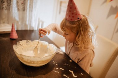 Parti şapkası takan genç bir çocuk kek hamuruyla dolu büyük bir kaseyi karıştırıyor. Renkli dekorasyonlar ve şenlikli bir atmosfer ailenin doğum günü kutlaması için neşeli bir ortam yaratır..