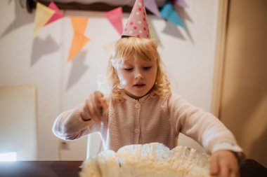 Şenlik şapkalı küçük bir çocuk aile toplantısında metal bir kasede kek hamuru karıştırıyor. Oda renkli afişlerle süslenmiş. Kutlama için neşeli bir ortam yaratıyor..