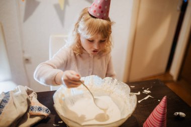 Genç bir kız büyük bir kasede kek hamuru karıştırıyor. Parti şapkası takarak mutfak masasındaki görevine odaklanıyor.. 