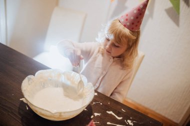 Konforlu bir ortamda, parti şapkalı genç bir kız aile kutlaması sırasında bir kasede kek hamuru karıştırıyor. Atmosfer sevinç ve heyecanla dolu. Kardeş bağı kurmak için mükemmel..