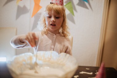 Parti şapkası takan küçük bir çocuk, evinde neşeli bir kutlama sırasında renkli süslemeler ve ailesiyle çevrili bir pastayı süslemeye odaklanıyor..