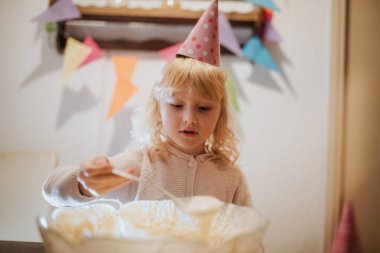 Parti şapkası takan genç bir çocuk, etrafı şenlikli süslemelerle çevriliyken kek hamurunu karıştırıyor. Ortam neşeli, özel bir aile kutlamasının altını çiziyorum..