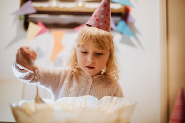Parti şapkalı küçük bir çocuk aile kutlaması sırasında kase içinde kek hamuru karıştırıyor. Renkli dekorasyonlar ve şenlikli bir atmosfer bu özel gün için neşeli bir hava yaratır..
