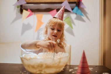 Genç bir kız parti şapkası takarken bir kase hamuru karıştırıyor. Renkli dekorasyonlar arka planda asılı, neşeli bir doğum günü ortamı yaratıyor.. 
