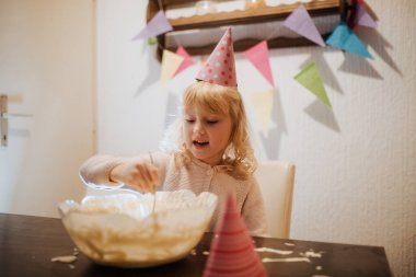 Genç bir kız bir aile kutlamasında bir kase kek hamurunu zevkle karıştırıyor. Renkli parti dekorasyonları odayı süslüyor, etrafında şenlikli bir atmosfer yaratıyor..
