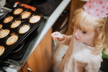 Parti şapkası takan genç bir çocuk sıcak bir mutfakta yeni pişmiş kekleri denetliyor ve heyecan gösteriyor.. 