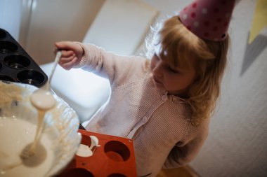 Küçük bir çocuk kaşık kullanarak, parti şapkası takarak hamuru karıştırmaya odaklanır. Mutfak aydınlık ve neşeli. Özel bir olayı kutlayan eğlenceli dekorasyonlar var..