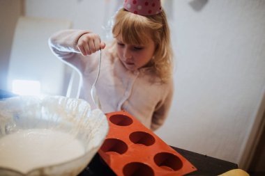 Genç bir çocuk kaşıkla hamuru karıştırıyor, silikon kalıbı doldurmaya hazırlanıyor. Parti şapkası takıyor ve sıcak bir mutfak ortamında pişirme sürecine odaklanıyor..