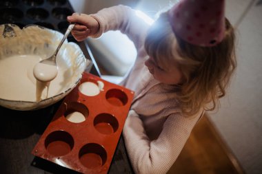 Küçük bir çocuk, parti şapkası takmış bir kaşıkla kek hamurunu karıştırıyor. Çocuk karışımı kırmızı kek kalıbına dökmeye odaklanmış. Aile neşesiyle dolu sıcak bir mutfak ortamında..