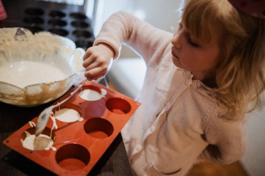 Küçük kız sıcak bir mutfakta neşeyle yemek pişiriyor. çocuk dikkatlice silikon kalıpları doldurur 