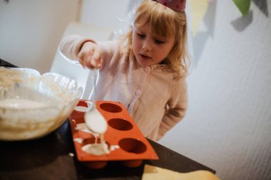 Genç bir çocuk, oyuncu bir parti şapkası takarken silikon pişirme kalıbının içine hamuru dökmeye odaklanıyor. Mutfak fırın aletleri ve dekorasyonlarla dolu. Sıcak bir atmosfer yaratıyor..