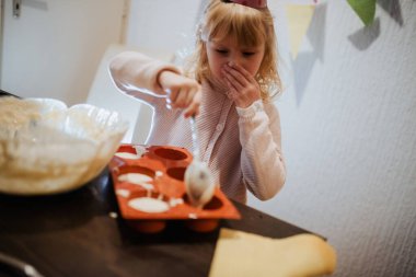 Neşeli bir mutfakta, kıvırcık saçlı genç bir kız ağzını şaşkınlıkla kapatırken hamuru dikkatlice bir tepsiye atar.. 