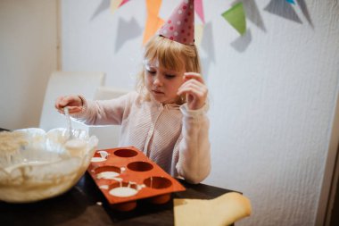 Parti şapkası takan genç bir çocuk evde ailesiyle kutlama yaparken kek tepsisine malzemeleri karıştırmakla meşgul. Renkli dekorasyonlar duvarı süsler, şenlikli atmosfere katar..