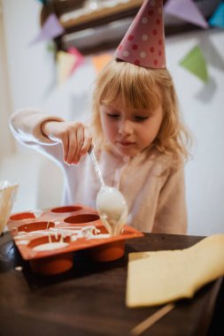 Genç bir kardeş, küçük bir masada ev yapımı ikramlar hazırlamaktan zevk alıyor. Parti şapkası takıyor, rahat bir yerde renkli dekorasyonlarla çevrili..