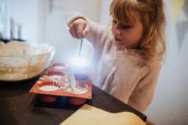 Küçük bir çocuk hafta sonu sabahı ailesiyle birlikte yemek pişirirken dikkatlice silikon kalıba hamur dökmektedir. Mutfakta pişirme malzemeleriyle dolu sıcak ve davetkar bir ortam var..