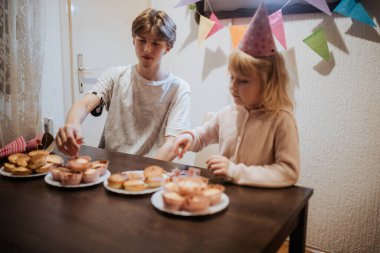 İki kardeş bir doğum günü partisinde kek süslemekle meşgul. Atmosfer şenlikli, renkli süslemeler ve kahkahalarla dolu, ortak sevinçlerini vurguluyorlar..