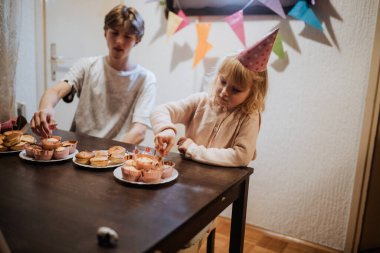 İki kardeş evde doğum günü kutlamalarının keyfini çıkarırken, kapkekleri mumlarla süslemeye odaklanmış, etrafı da şenlikli süslemeler ve ikramlar ile çevrili..