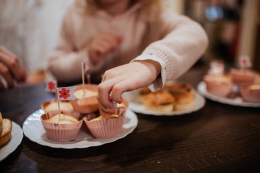 İki kardeş, üzerinde bayraklar olan renkli çöreklerle dolu bir tatlı masasının etrafında toplanıyorlar. Evde neşeli bir aile toplantısı sırasında tatlı ikramlardan hoşlanırlar..