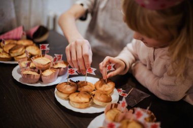 İki kardeş bir aile toplantısında mutlu bir şekilde kek süslüyor. Lezzetli ikramlarla ve sıcak, misafirperver bir atmosferle çevriliyken küçük bayraklar ekliyorlar..