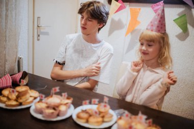 İki kardeş, doğum günlerini kutlarken renkli süslemeler ve kapkek tabakları ile çevrili olarak evlerinde neşeli bir anın tadını çıkarıyorlar. Atmosfer neşeyle dolu..