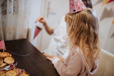 Parti şapkalı küçük bir çocuk, tatlı ikram ve süslemelerle dolu bir masada oturmuş, bir aile üyesiyle eğlenirken aynı zamanda kahkahalarını paylaşıyor..