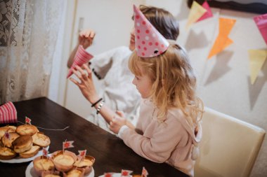 İki kardeş, renkli parti şapkaları takarak ve kek dolu bir masada oturarak neşeli bir atmosfer ve tatlı anılar yaratarak evde eğlenceli bir kutlamanın tadını çıkarıyorlar..