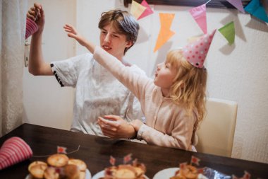 İki kardeş, parti şapkaları takıp ikramda bulunmak için evde toplanmaktan hoşlanıyorlar. Renkli dekorasyonlar kutlamaları sırasında neşeli bir atmosfer yaratıyor..