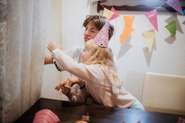 İki kardeş, renkli süslemelerle dolu bir doğum günü kutlamasında neşeyle etkileşime geçiyorlar. Heyecan ve coşku dolu, neşeli bir andalar..