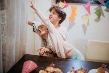 İki kardeş, şenlikli bir toplantıda neşeli bir anı paylaşıyor. Şekerler ve renkli afişlerle süslenmiş bir masanın yanında şakacı bir şekilde etkileşime geçerek sıcak bir aile ortamı yaratıyorlar..