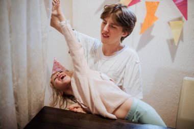 Kardeşler, renkli süslerle çevrili ve parti şapkaları takmış bir doğum günü kutlaması sırasında evde keyifli bir anın tadını çıkarırlar. Mutluluk birlikte çalarken havayı doldurur..