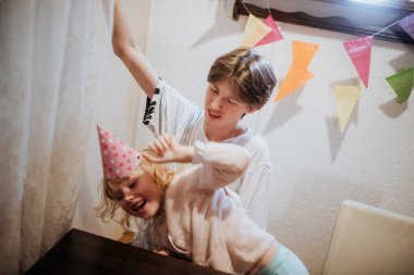 İki kardeş renkli süslemelerle süslenmiş bir doğum günü kutlamasının tadını çıkarıyorlar. Kahkahalar havayı dolduruyor, neşeli bir anı paylaşıyorlar, parti şapkaları takıp eğleniyorlar..