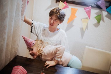 Evde bir doğum günü kutlaması sırasında iki kardeş arasında neşeli bir etkileşim başlar. Büyük kardeş küçük olana parti şapkası ile yardım ederken renkli süslemeler odayı aydınlatıyor..