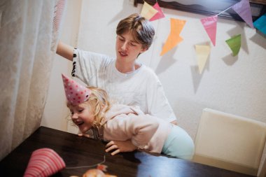 İki kardeş, canlı dekorasyonlarla çevrili bir doğum günü kutlamasında aynı kahkahayı paylaşıyor. Büyük kardeş neşeli bir şekilde etkileşime girerken küçük kardeş neşeli atmosferin tadını çıkarıyor..