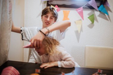 Neşeli bir an, küçük bir çocuğun doğum günü kutlamasında parti şapkası takmasına neşe içinde yardım eden bir kardeş olarak gözler önüne seriliyor. Renkli dekorasyonlar rahat odayı dolduruyor, şenlikli atmosferi arttırıyor..