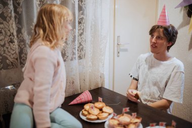 İki kardeş, evde parti şapkaları takıp kek paylaşarak neşeli bir anın tadını çıkarıyorlar. Atmosfer neşeli, kahkahalarla ve kardeş bağlarıyla dolu..